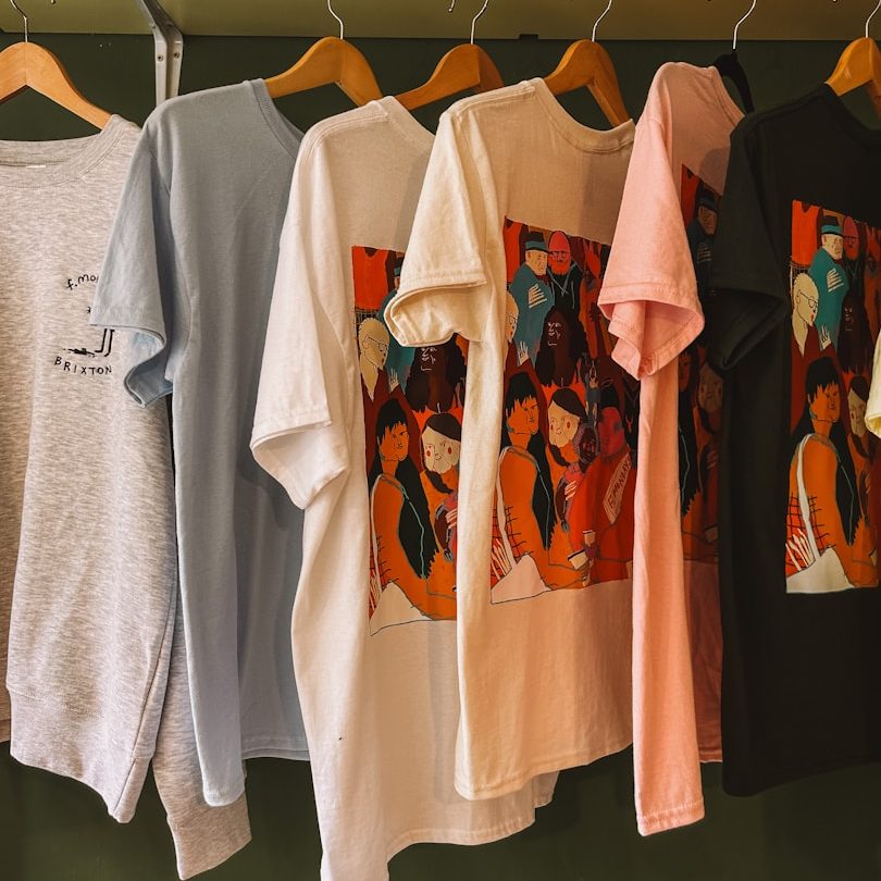 Textildruck & Werbemittel Sechs T-Shirts mit farbigen Grafiken hängen an Holzbügeln.